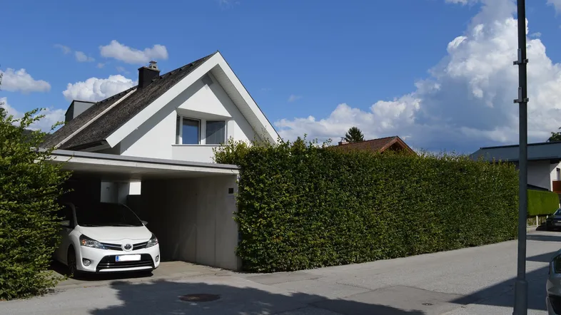 Weißes Einfamilienhaus mit Satteldach, Carport und hoher Hecke an der Straße.