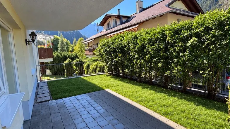 Gepflasterte Terrasse mit Rasenfläche und Hecke, umgeben von grüner Landschaft und Bergen.