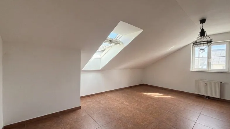 Helles Dachgeschosszimmer mit Dachschräge, Fliesenboden und einem großen Oberlicht.