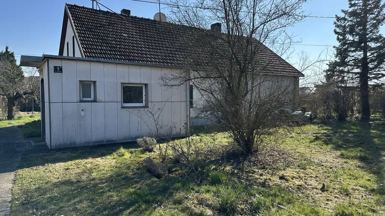 Außenansicht eines Hauses mit Anbau und ungepflegtem Gartenbereich unter blauem Himmel.