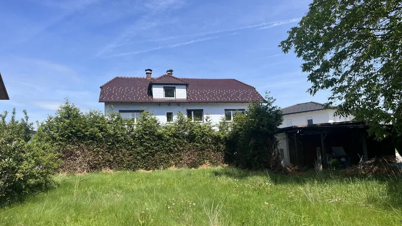 Weißes Haus mit rotem Dach und großem, ungemähtem Gartenbereich unter blauem Himmel.