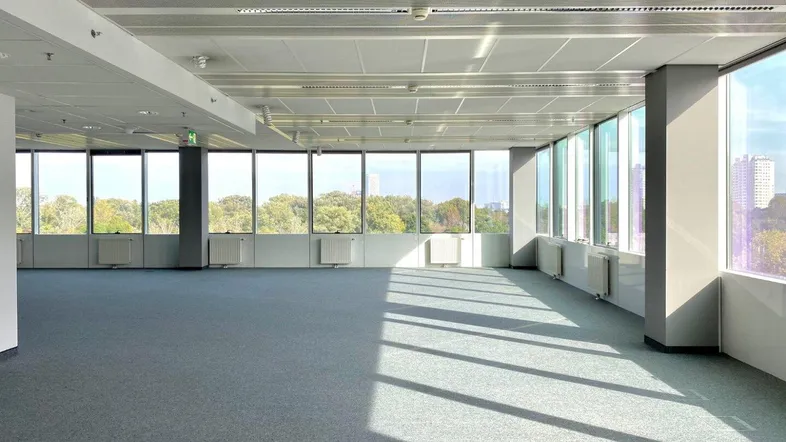 Großer, leerer Büroraum mit Teppichboden und raumhohen Fenstern, die viel Tageslicht hereinlassen.