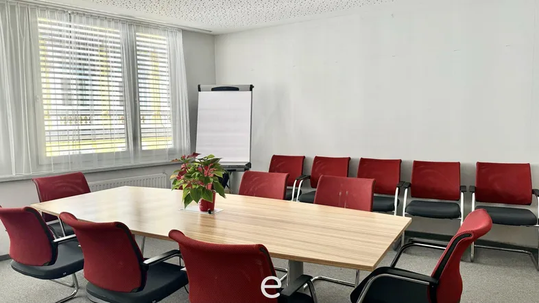 Heller Konferenzraum mit großem Holztisch, roten Stühlen und einem Whiteboard für Besprechungen.