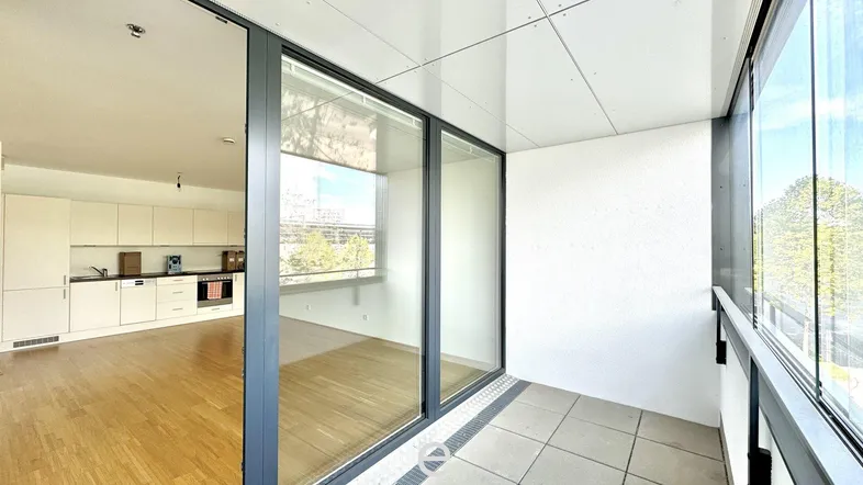 Offener Wohnbereich mit integrierter weißer Küche, Holzboden und Zugang zu einer verglasten Loggia.