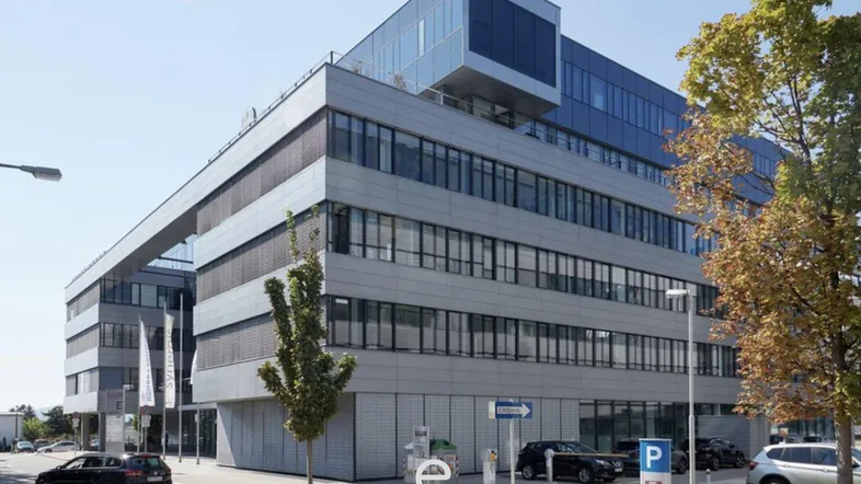 Modernes Bürogebäude mit Glasfassade, Parkplätzen und Ladestationen unter blauem Himmel.