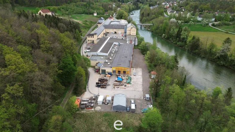Luftaufnahme eines großen Industrie- oder Gewerbekomplexes am Flussufer, umgeben von viel Grün und Bäumen.