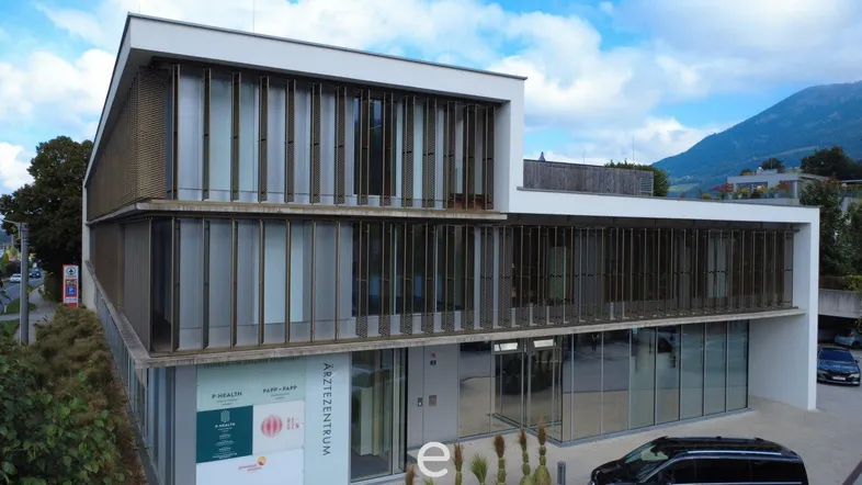 Modernes Ärztezentrum mit großer Glasfassade und Lamellen an der Außenseite, umgeben von Natur und Bergen.