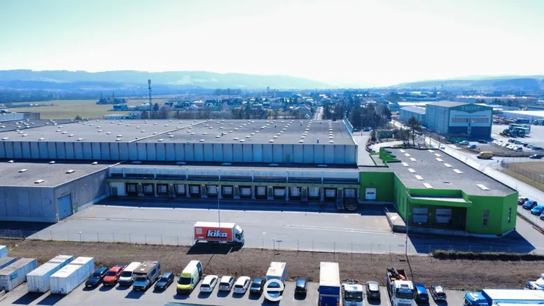 Luftaufnahme eines großen Industrie- und Logistikzentrums mit Laderampen und Parkflächen, umgeben von ländlicher Landschaft und Stadtblick.