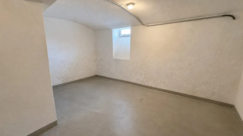 Heller Kellerraum mit Betonboden, kleinem Fenster und sichtbaren Leitungen an der Decke.