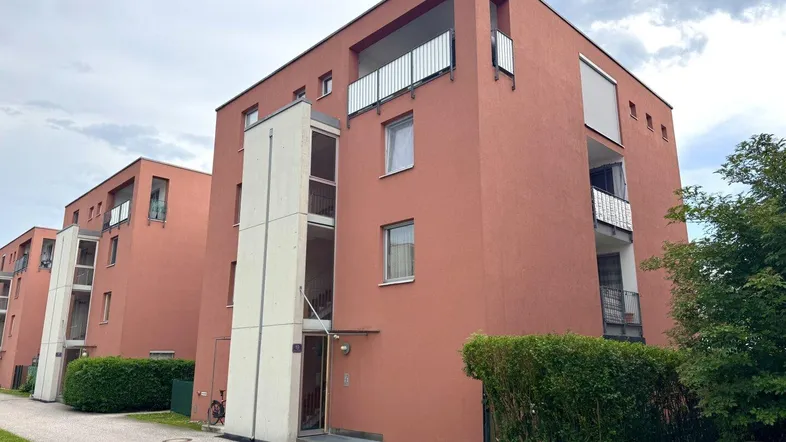 Modernes Mehrfamilienhaus mit roter Fassade und Balkonen unter bewölktem Himmel.