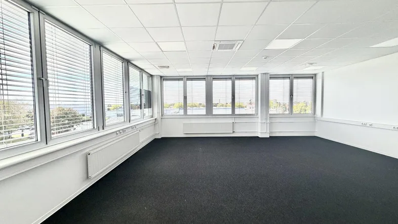 Geräumiges, leeres Büro mit dunklem Teppichboden, vielen Fenstern und Jalousien, bereit zur Nutzung.