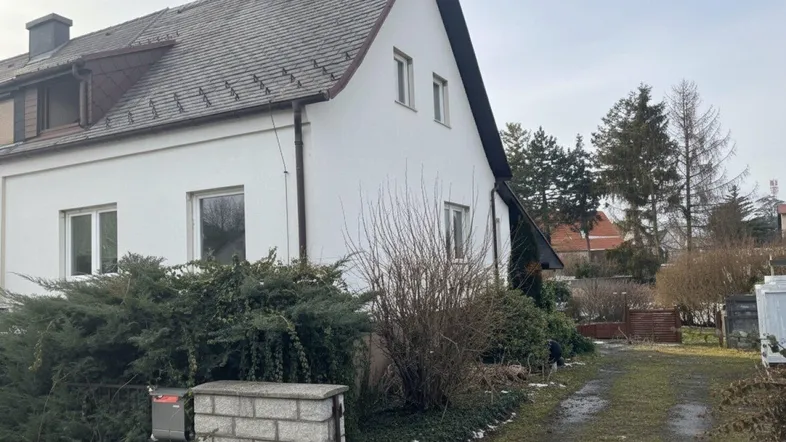 Weißes Einfamilienhaus mit Satteldach, umgeben von Sträuchern und einem Vorgarten.