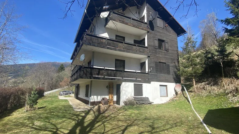 Traditionelles Holzhaus mit mehreren Balkonen und Bergblick, umgeben von grüner Wiese unter blauem Himmel.