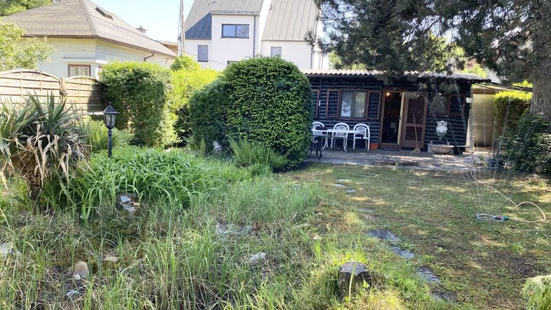 Garten mit Rasenfläche, einer kleinen Holzhütte und Sitzgelegenheiten im Freien, im Hintergrund ein Wohnhaus.