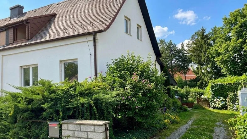Seitenansicht des Hauses mit Kiesauffahrt und üppigem Grün im Vorgarten bei Sonnenschein.