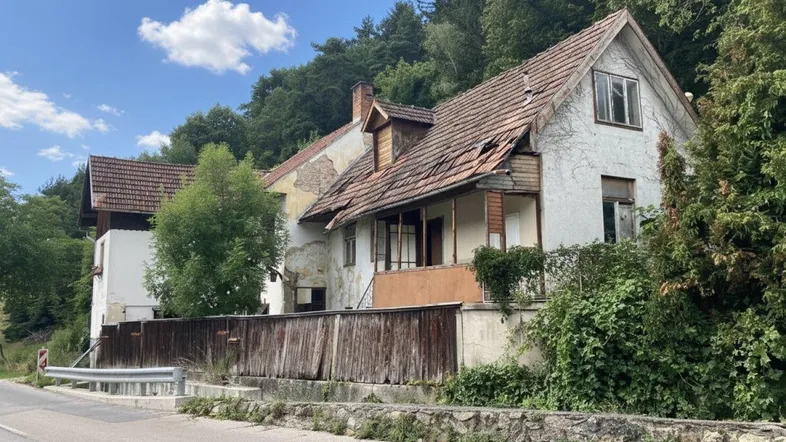 Altes, baufälliges Haus mit beschädigtem Dach und Fassade, umgeben von Bäumen und einer Straße.