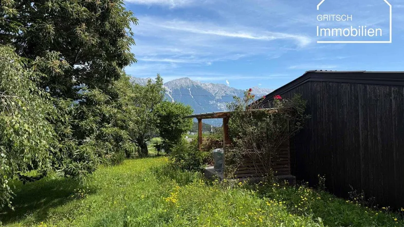 Blick auf das Gebäude von einem grünen Gartenbereich mit Bäumen und Bergen im Hintergrund.