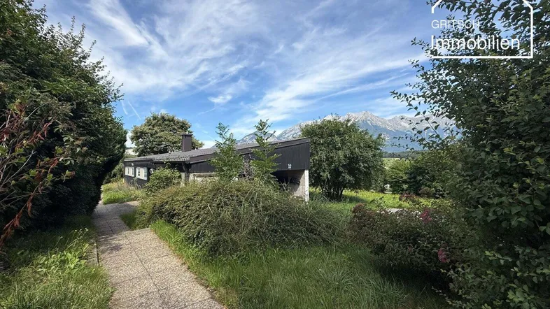 Einfamilienhaus mit üppigem Garten und malerischem Bergblick unter blauem Himmel.
