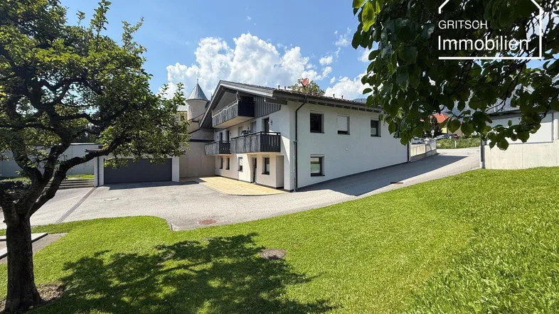 Modernes Wohnhaus mit Balkonen, umgeben von einer grünen Rasenfläche und Bäumen unter blauem Himmel.