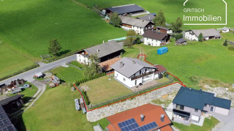 Luftaufnahme eines Hauses mit Grundstück in ländlicher Umgebung, umgeben von grünen Wiesen und Bergen.