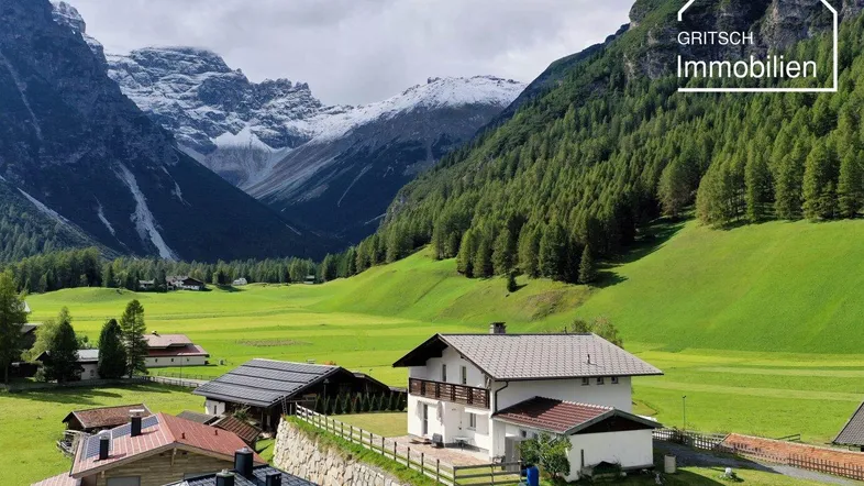 Malerisches Haus in einem grünen Tal mit schneebedeckten Bergen im Hintergrund und viel Natur.