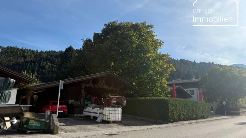 Überdachter Stellplatz und Zufahrt zu einem Gebäude mit grüner Hecke und Bergkulisse.