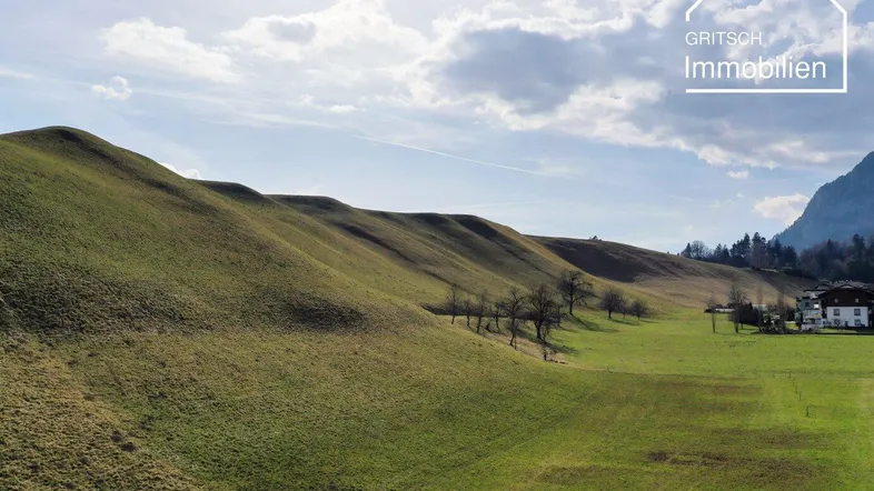 Weitläufige grüne Hügellandschaft mit einem Haus in der Ferne und Bergblick unter bewölktem Himmel.