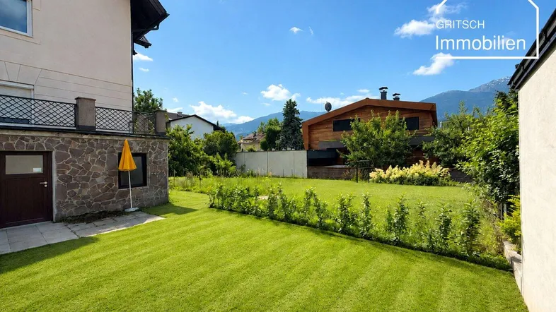 Gepflegter Garten mit Rasenfläche, Hecke und Bergblick unter blauem Himmel.