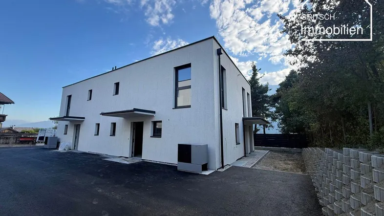 Moderne weiße Fassade eines Neubaus mit klaren Linien und dunklen Fensterrahmen unter blauem Himmel.