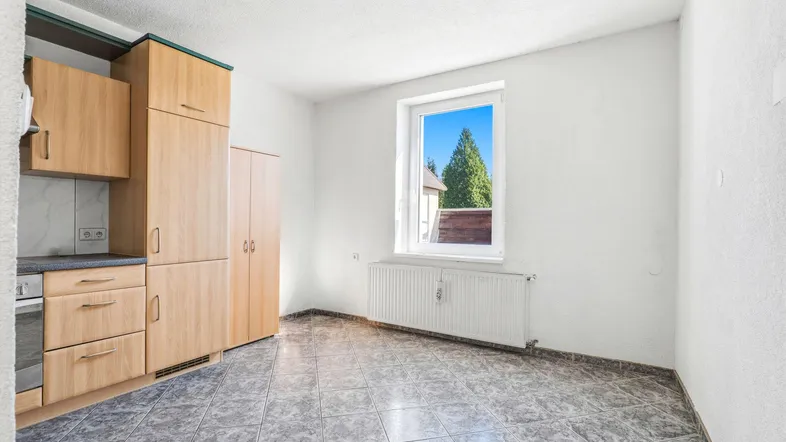 Helle Küche mit Holzschränken, Fliesenboden und Blick ins Grüne durch das Fenster.