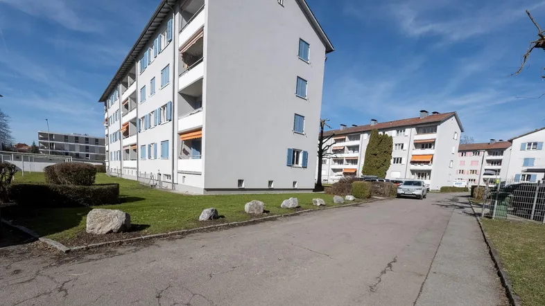 Mehrfamilienhaus mit gepflegter Grünfläche, blauen Fensterläden und weiteren Wohngebäuden in der Umgebung.