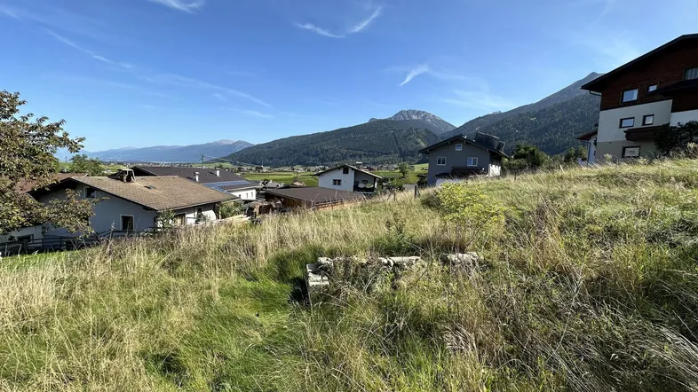 Blick über ein sonniges Grundstück mit hohem Gras auf umliegende Häuser und eine beeindruckende Berglandschaft.