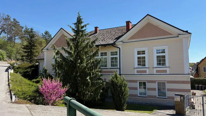 Traditionelles Haus mit heller Fassade, markanten Giebeln und umgebender Grünfläche an einem sonnigen Tag.