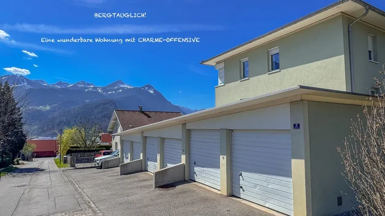 Mehrfamilienhaus mit Garagen und beeindruckendem Bergblick im Hintergrund unter blauem Himmel.