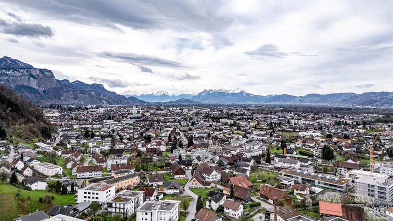 Panoramablick über eine Stadt mit schneebedeckten Bergen im Hintergrund unter bewölktem Himmel.