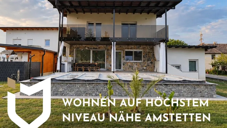 Modernes Haus mit Steinfassade, Balkon und Terrasse, umgeben von Grünflächen unter blauem Himmel.