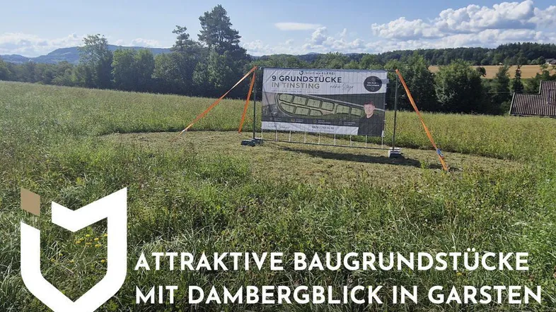 Grünes Feld mit einem Schild, das Baugrundstücke bewirbt, umgeben von einer ländlichen Landschaft mit Hügeln und blauem Himmel.