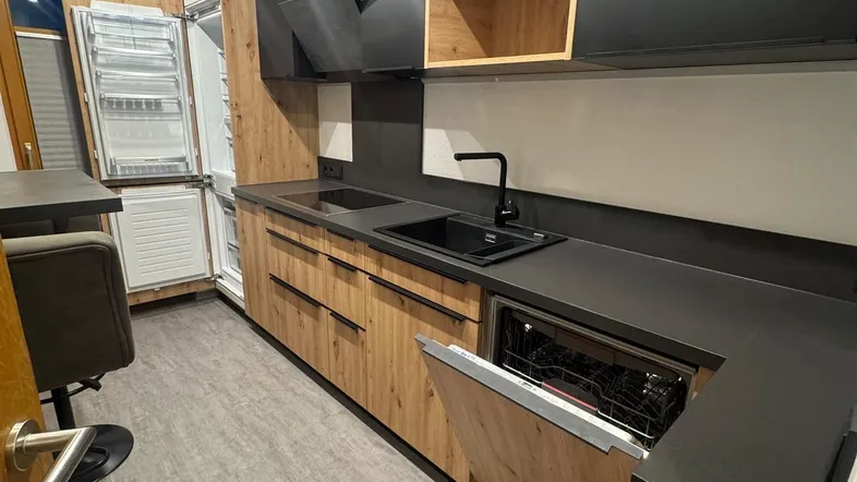 Moderne Küche mit Holzfronten, dunkler Arbeitsplatte und integrierten Geräten sowie Kühlschrank.