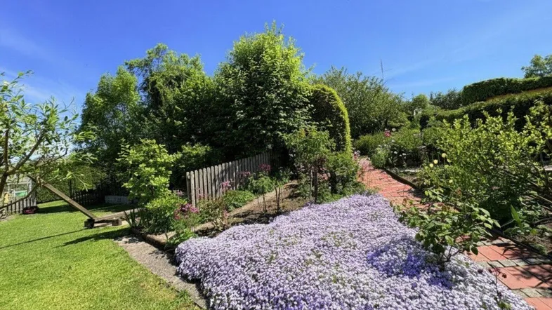 Grüner Garten mit blühenden Blumenbeeten, Rasenfläche und einem kleinen Spielbereich unter blauem Himmel.