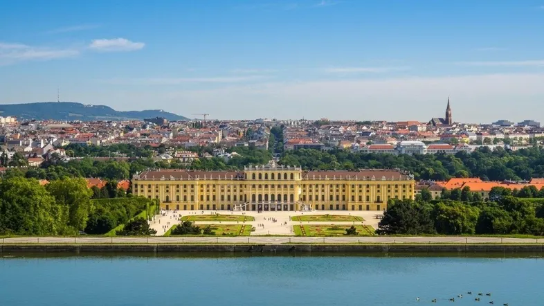 Panoramablick auf ein historisches Schloss mit Wasserfläche und Stadtkulisse im Hintergrund unter blauem Himmel.