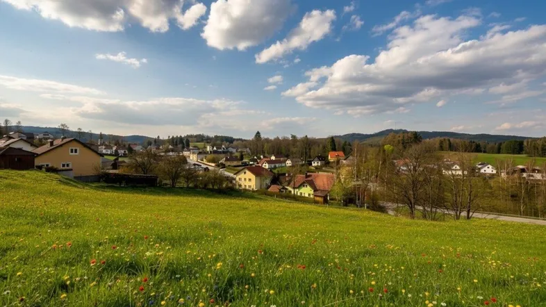 Weitläufige Landschaft mit einem malerischen Dorf, umgeben von grünen Wiesen und sanften Hügeln unter blauem Himmel.