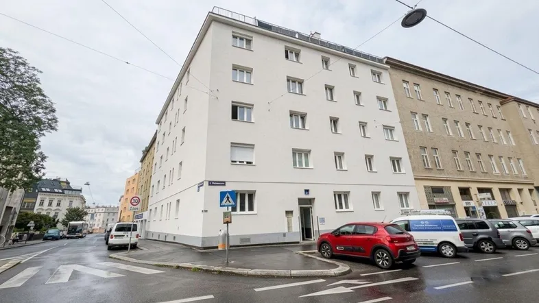 Weißes Mehrfamilienhaus an einer Straßenecke mit parkenden Autos davor.