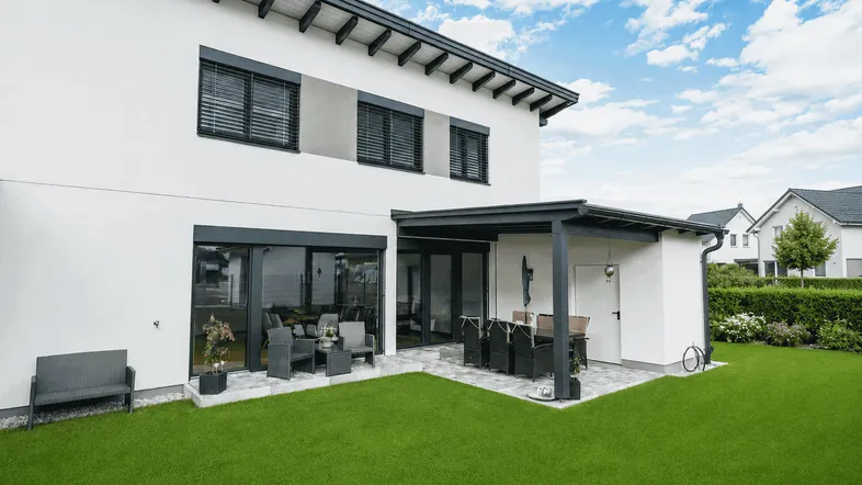 Moderne Hausrückseite mit großer Rasenfläche, überdachter Terrasse und großen Glasfronten.