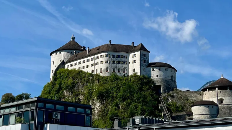 Historische Festung auf einem bewaldeten Hügel mit modernen Gebäuden im Vordergrund unter blauem Himmel.