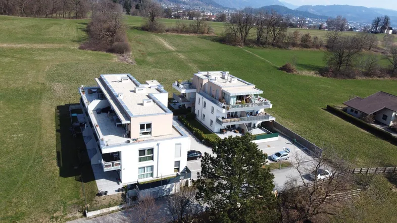 Luftaufnahme von modernen Wohngebäuden in einer grünen Landschaft mit Bergen im Hintergrund.