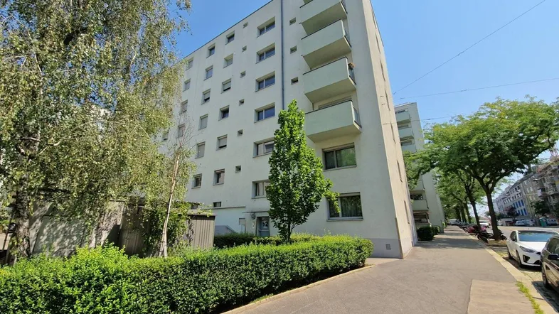 Mehrstöckiges Wohngebäude mit Balkonen und Bäumen an einer sonnigen Straße.