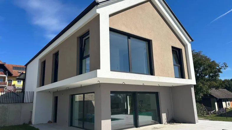 Modernes Einfamilienhaus mit großen Fenstern, heller Fassade und einer gepflasterten Terrasse.