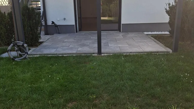 Überdachte Terrasse mit grauen Fliesen und angrenzendem Rasenbereich hinter dem Haus.