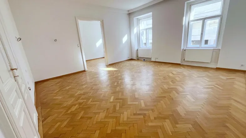 Geräumiges Wohnzimmer mit Fischgrätparkett und großen Fenstern, die viel Tageslicht hereinlassen.