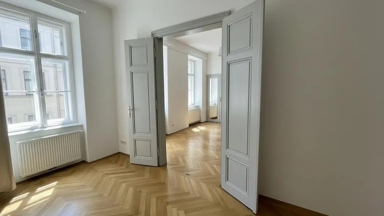 Helles Zimmer mit Fischgrätparkettboden, hohen Decken und großen Fenstern, das zu einem weiteren Raum führt.